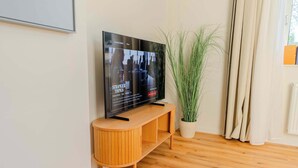 Litteänäyttöinen televisio puisen jalustan päällä, ruukussa oleva kasvi ja ikkuna, jossa on verhot.