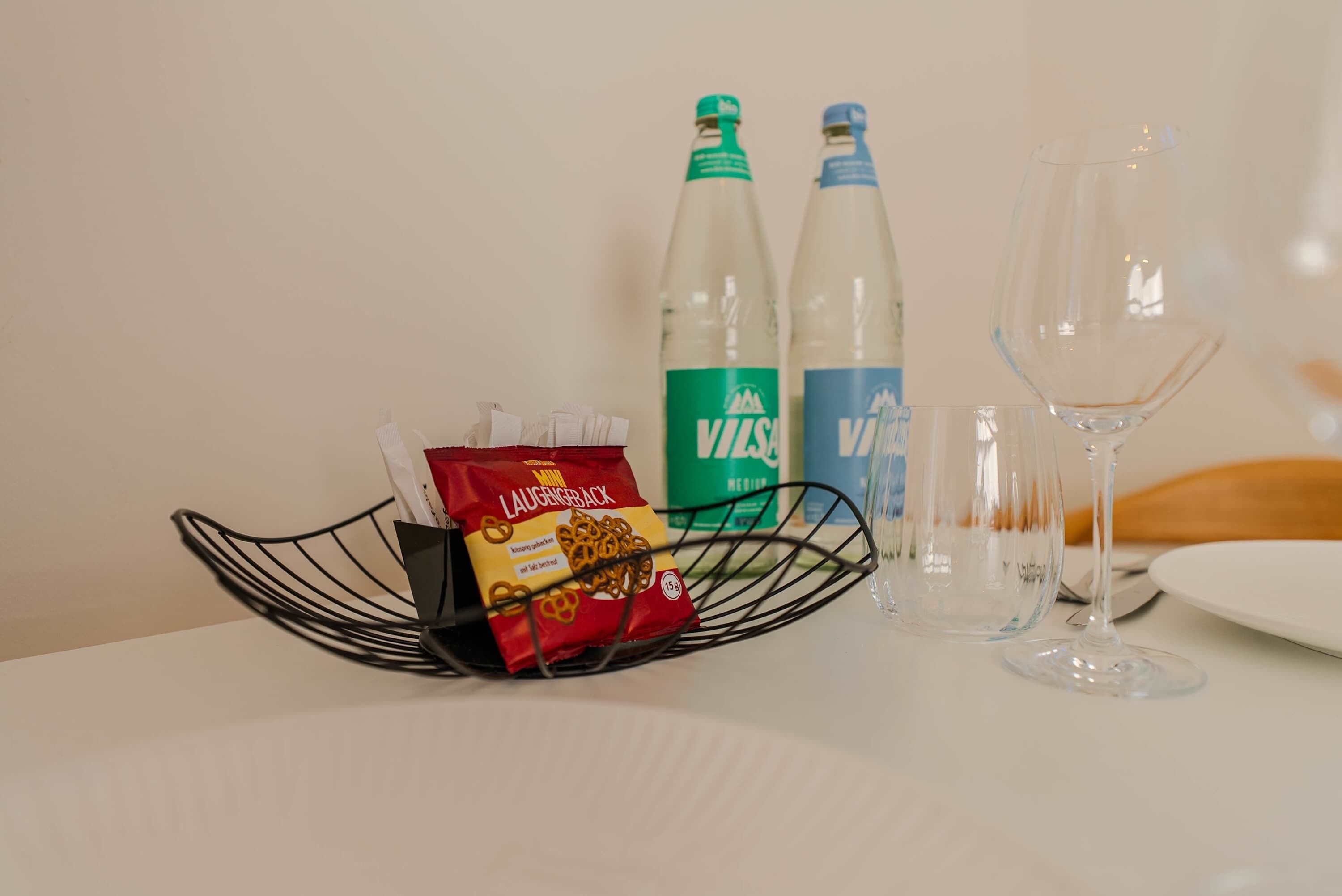 Et bord med en wirekurv, der indeholder snacks, to flasker vand og to tomme vinglas.