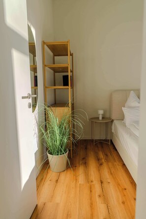 Une chambre avec un plancher en bois, un lit, une table de chevet avec une lampe, une bibliothèque et une plante en pot.