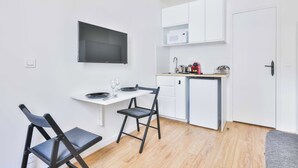Une cuisine compacte avec des meubles blancs, un four à micro-ondes et un petit plan de travail. On y trouve une table à manger avec des chaises et un téléviseur à écran plat fixé au mur.