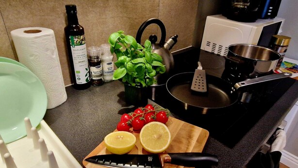 Un piano di lavoro in cucina con un tagliere, un coltello, pomodori, fettine di limone, una bottiglia d'olio d'oliva, un flacone di pepe nero, un contenitore di sale grosso, una pianta di basilico, un bollitore, una grattugia e un rotolo di carta assorbente.