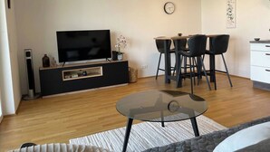 Un salon moderne avec un téléviseur à écran plat, une table basse en verre, un coin repas avec une table et des chaises, un coin cuisine avec un meuble blanc et une horloge au mur.