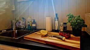 Uma bancada de cozinha com tábua de corte, faca, limão e alguns tomates. Há também frascos de produtos de limpeza e um vaso com planta.