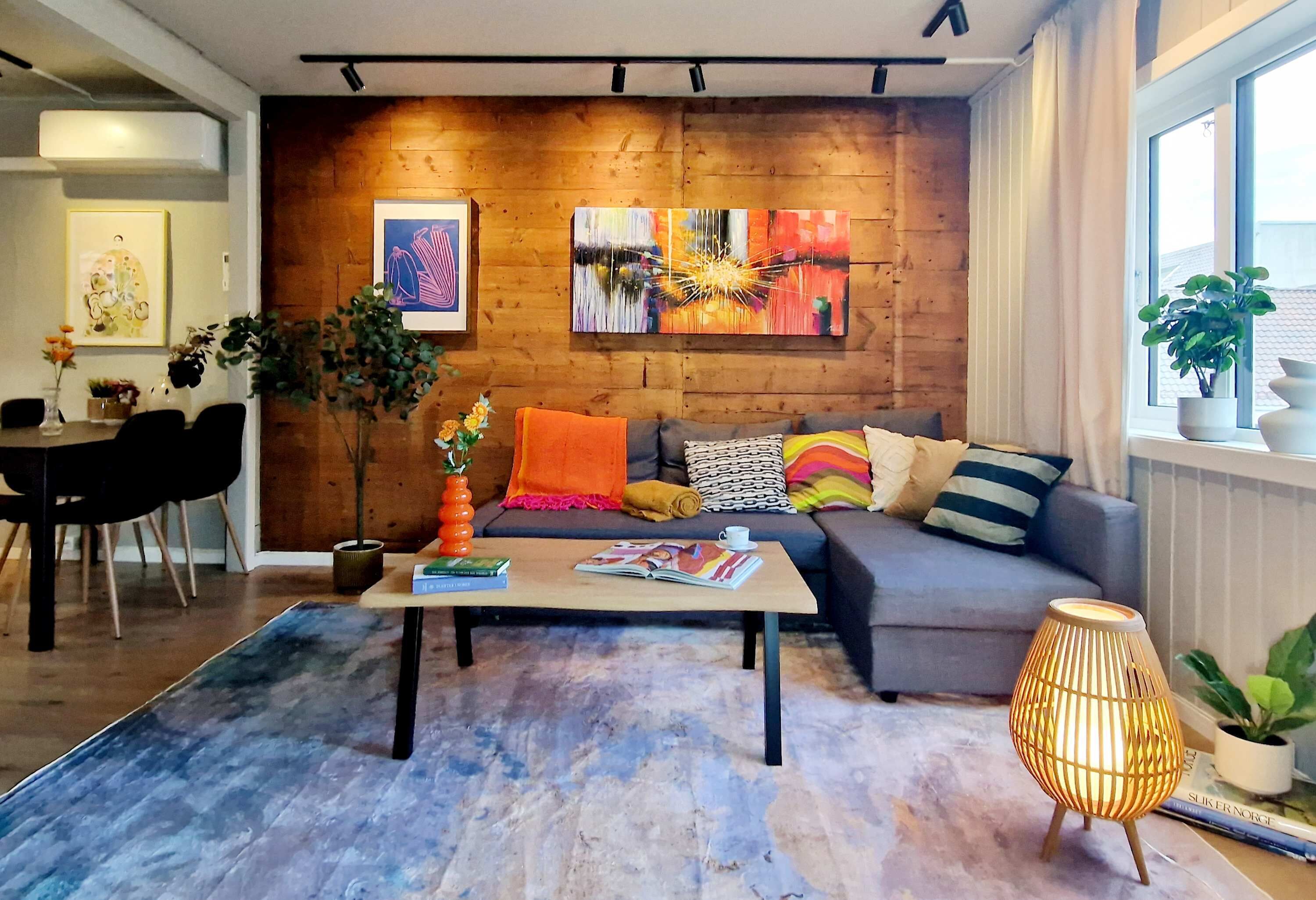 A modern living room with a wooden accent wall, a sofa with colorful cushions, a coffee table with books, and a floor lamp.