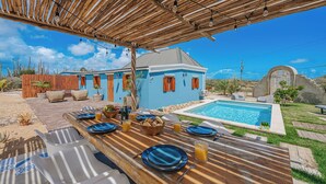 Un espace extérieur aménagé pour les repas, avec une piscine, une pergola en bois et une maison bleue en arrière-plan.
