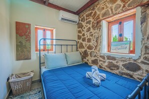 A bedroom with a stone wall, a bed with blue bedding, a metal headboard, a wicker basket labeled 'HOE', and a painting of a boat.
