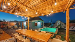 Une terrasse extérieure avec une table et des chaises en bois, une piscine et une maison en arrière-plan.