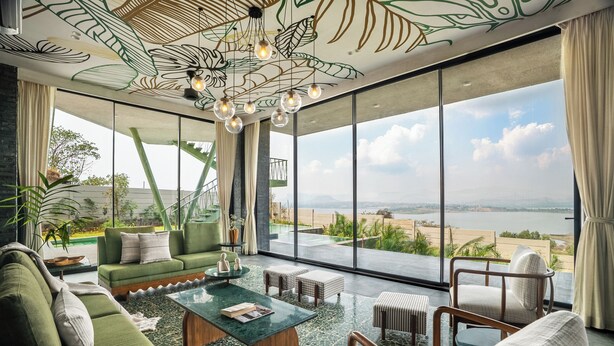 A spacious living room with a green sofa, a coffee table, and a view of a beach and ocean through large windows. - Verdantia (Igatpuri)
