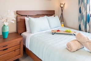 Ein ordentlich bezogenes Bett mit einer hellblauen Tagesdecke, ein Nachttisch mit einer Vase und einer Lampe, und ein Tablett mit Früchten und einem Glas Saft auf dem Bett.