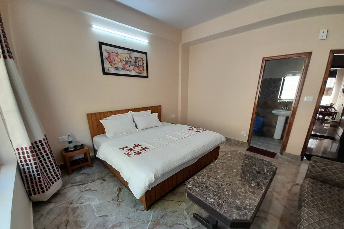 A bedroom with a bed, bedside table, and a small nightstand. There is a bathroom visible through an open door.