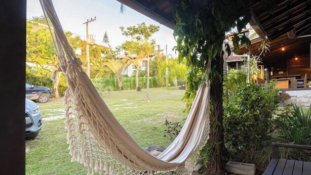 A hammock hanging over a grassy area with a building in the background. - Casas Valley Design - Praia do Rosa - SC (Imbituba)