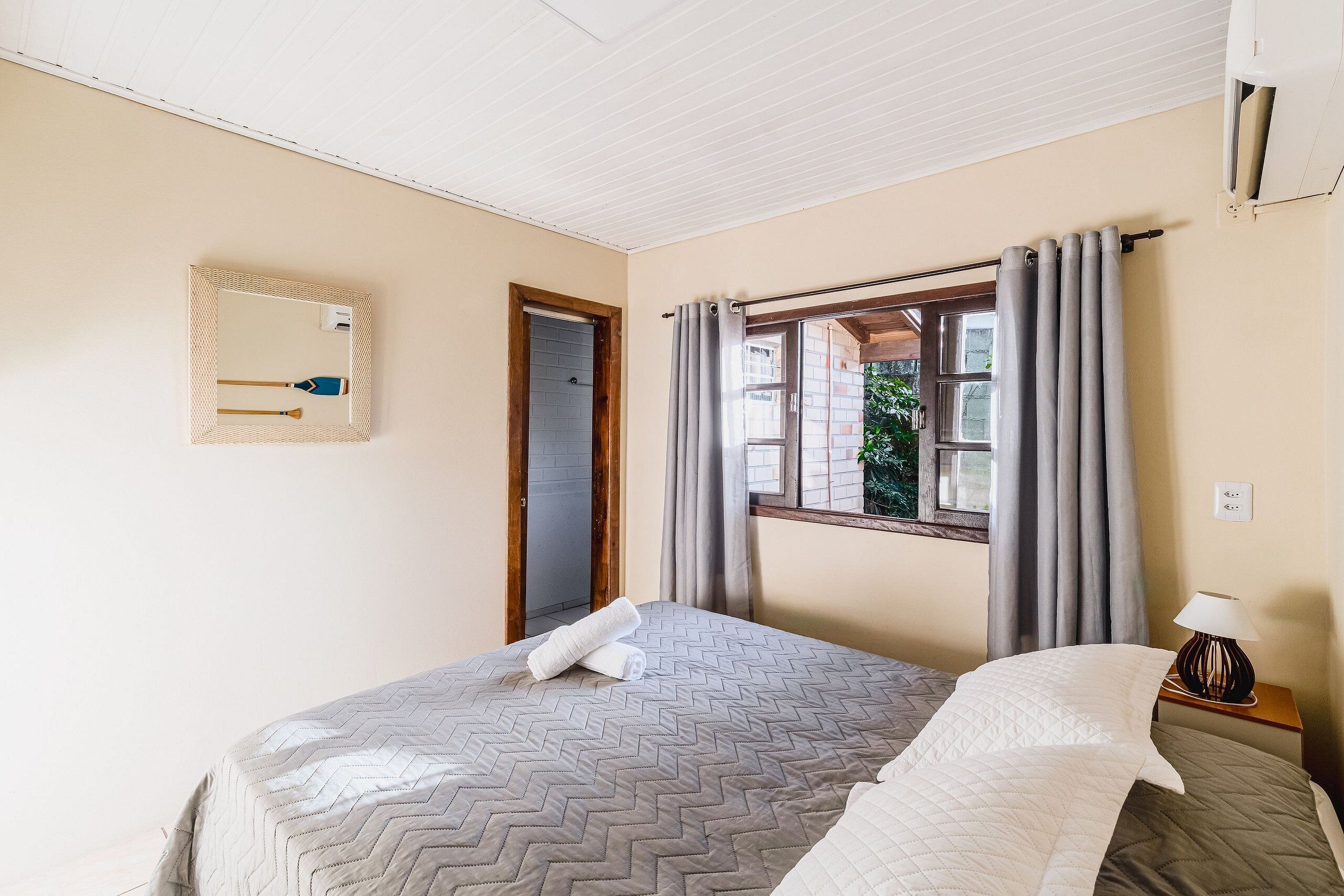 A bedroom with a bed, a bedside table with a lamp, a window with curtains, and a mirror on the wall.