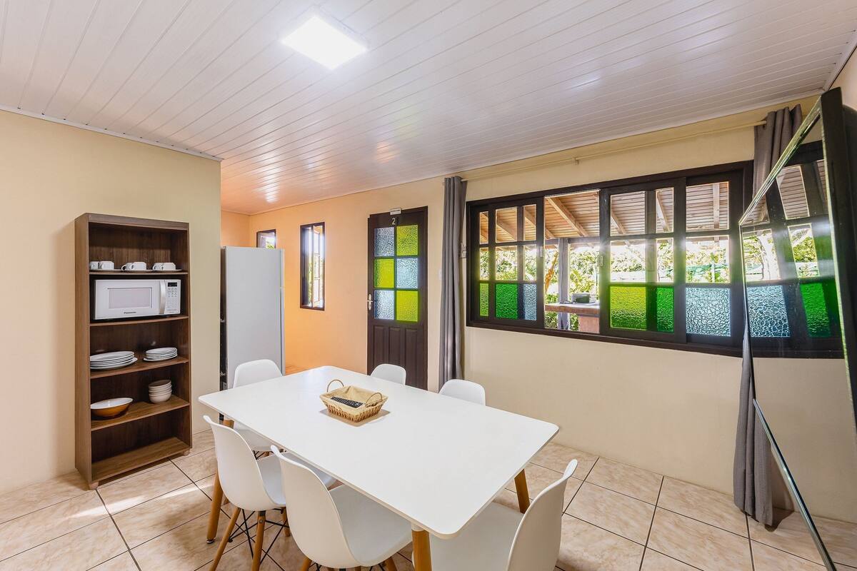 A room with a white table, white chairs, a wooden shelf with kitchenware, a refrigerator, and a window with a view of greenery.