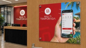 A hotel reception area with a wooden reception desk, a red wall with the hotel's logo, and a sign featuring a smartphone displaying a booking interface. - Casas Valley Design - Praia do Rosa - SC (Imbituba)
