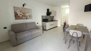 Un saló modern amb un sofà gris, una taula de dinar amb caires, una zona de cuina amb microones i armaris, i un quadre a la paret.