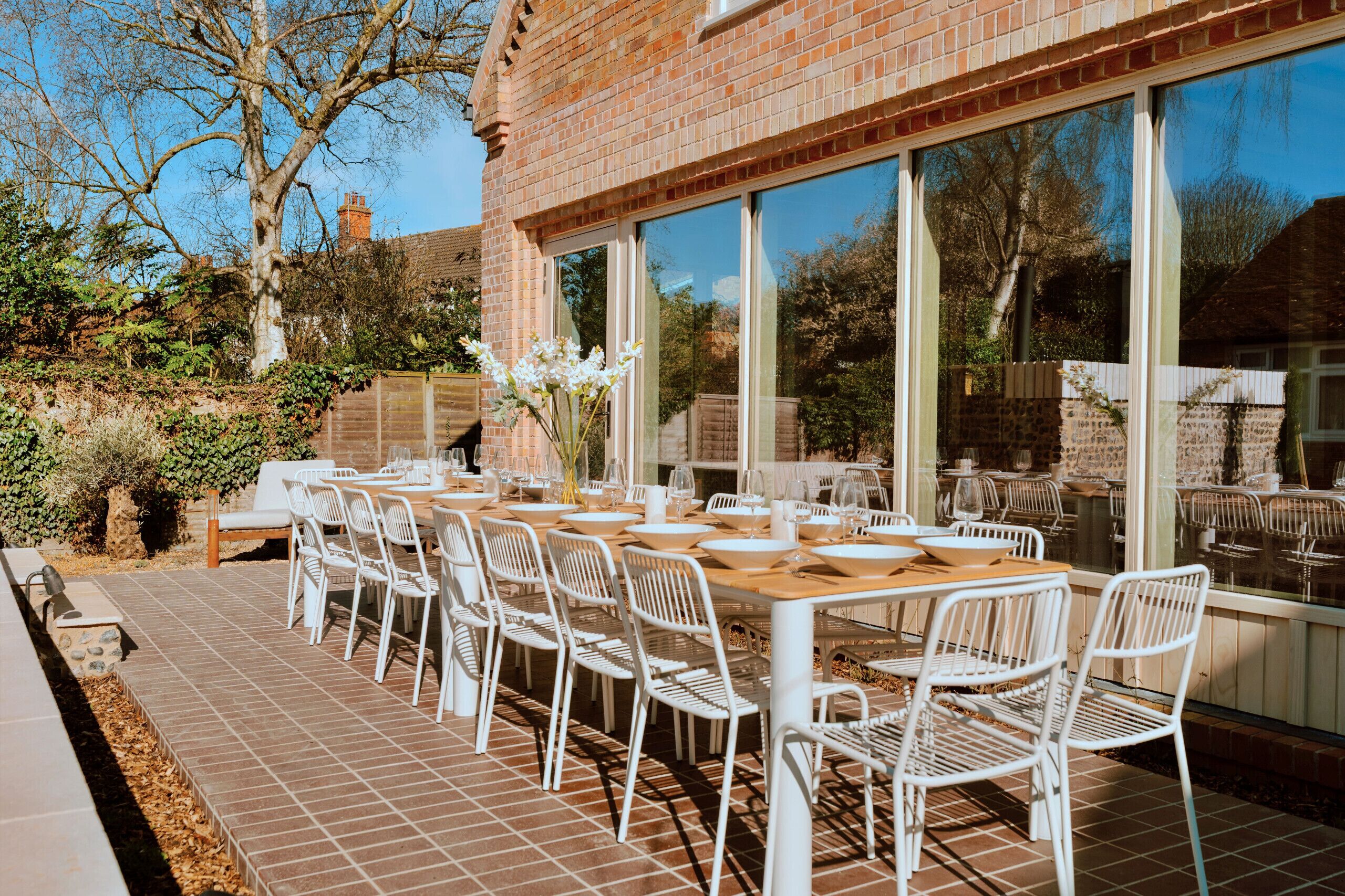 Un área de comedor al aire libre con un edificio de ladrillo, ventanas grandes y sillas de metal blanco.