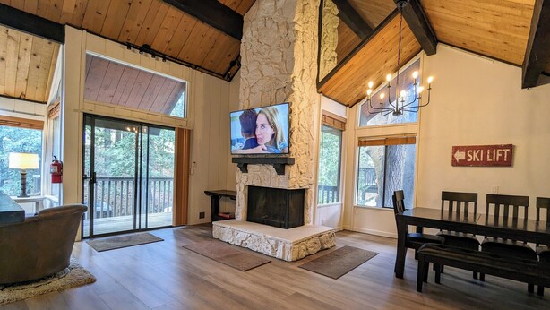 A spacious living room with a stone fireplace, wooden beams, a television, and a sign indicating a ski lift. - Snow Summit Retreat with Fireplace and Stunning Views (Big Bear Lake)