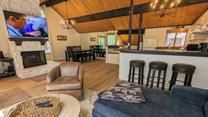 A cozy living room with a stone fireplace, leather armchair, and a blue sofa. A kitchen area with bar stools is visible in the background. - Snow Summit Retreat with Fireplace and Stunning Views (Big Bear Lake)