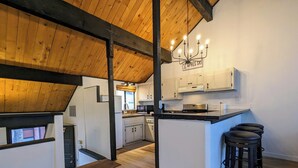A modern kitchen with wooden beams, white cabinets, a central island with stools, and a chandelier. - Snow Summit Retreat with Fireplace and Stunning Views (Big Bear Lake)