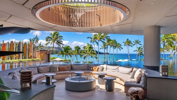Un área lounge moderna con una disposición de asientos curvada, una fogata en el centro y vistas al océano y palmeras.