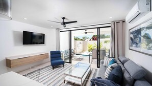 Un salon moderne doté d’un téléviseur à écran plat, d’une table basse en verre, d’un canapé, d’un fauteuil bleu et d’une porte-fenêtre coulissante donnant sur un espace extérieur avec une piscine.