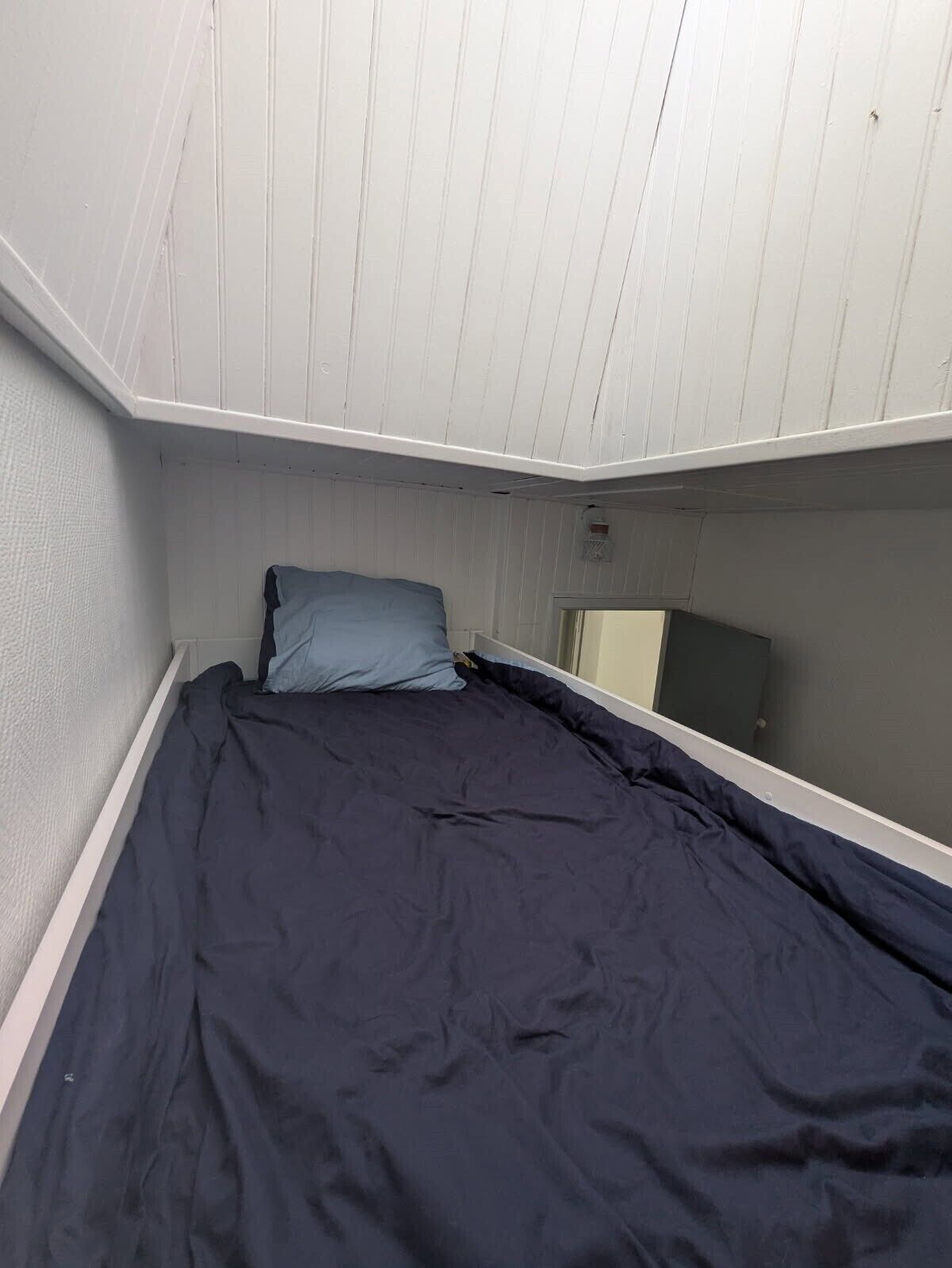 Een kleine, smalle kamer met een enkel bed, een raam en een spiegel aan de muur.