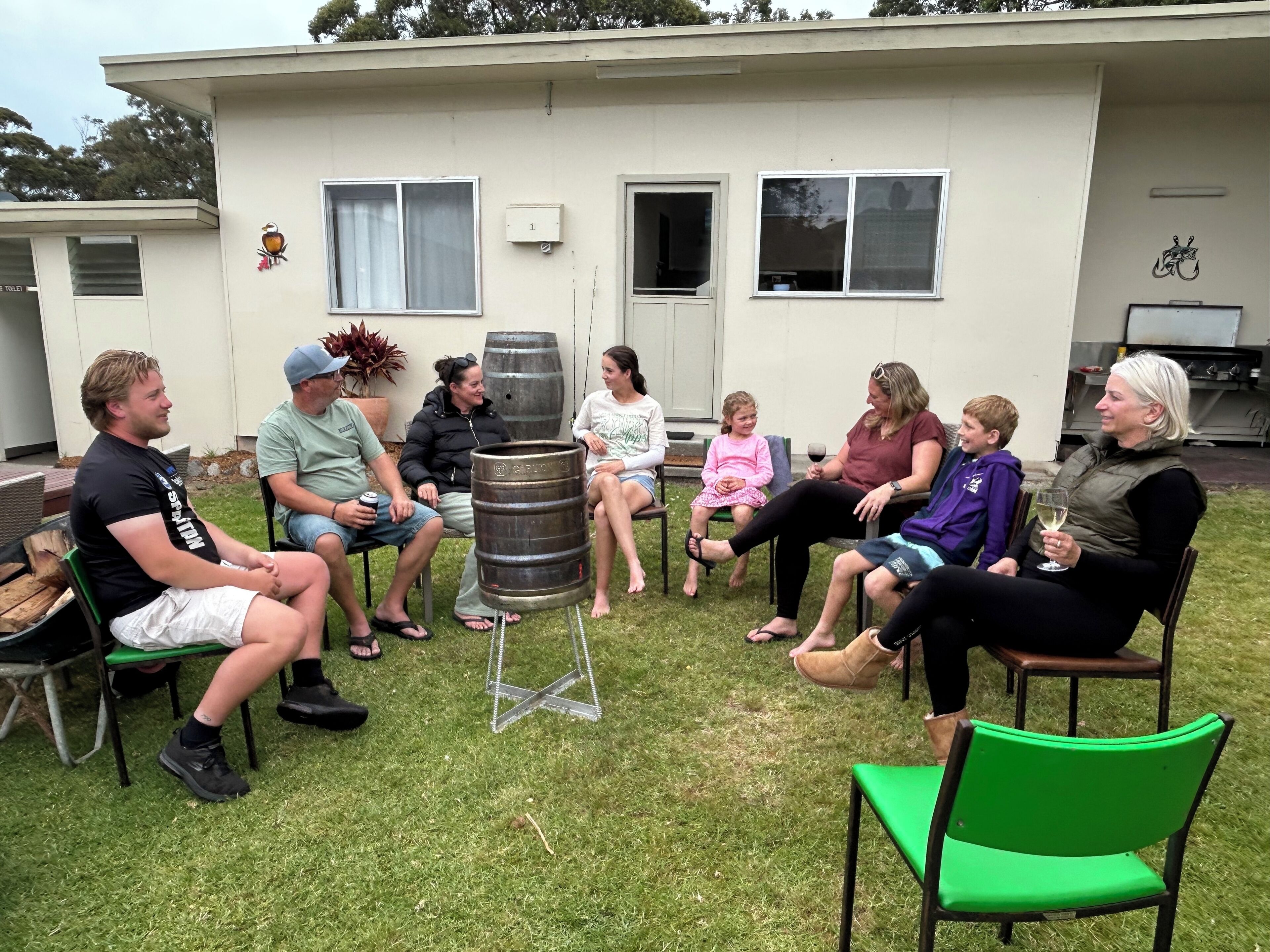 A group of people seated outdoors in a semi-circular arrangement around a central object, possibly a barrel, with a building in the background.