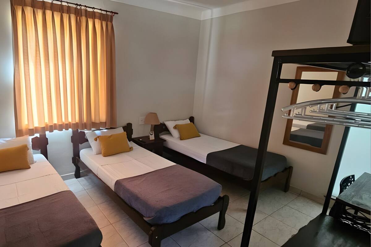 A hotel room with two single beds, a bunk bed, a window with curtains, a bedside table with a lamp, and a mirror.