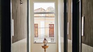 A hallway with a wooden floor, a window with a view of buildings, and a decorative vase on a stand. - Hotel Barbacena (Barbacena)