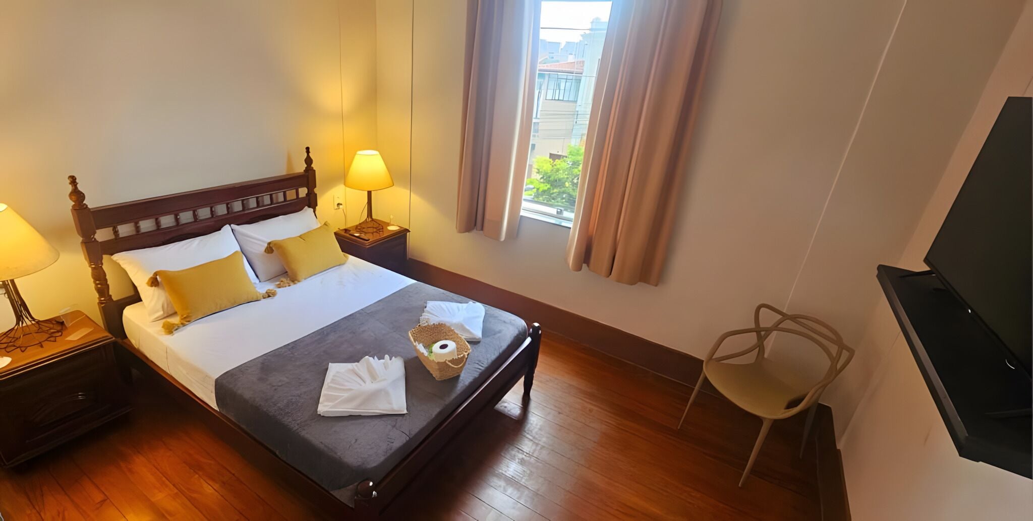 Une chambre d’hôtel avec un lit en bois, deux lampes de chevet, un téléviseur à écran plat et une fenêtre avec des rideaux.