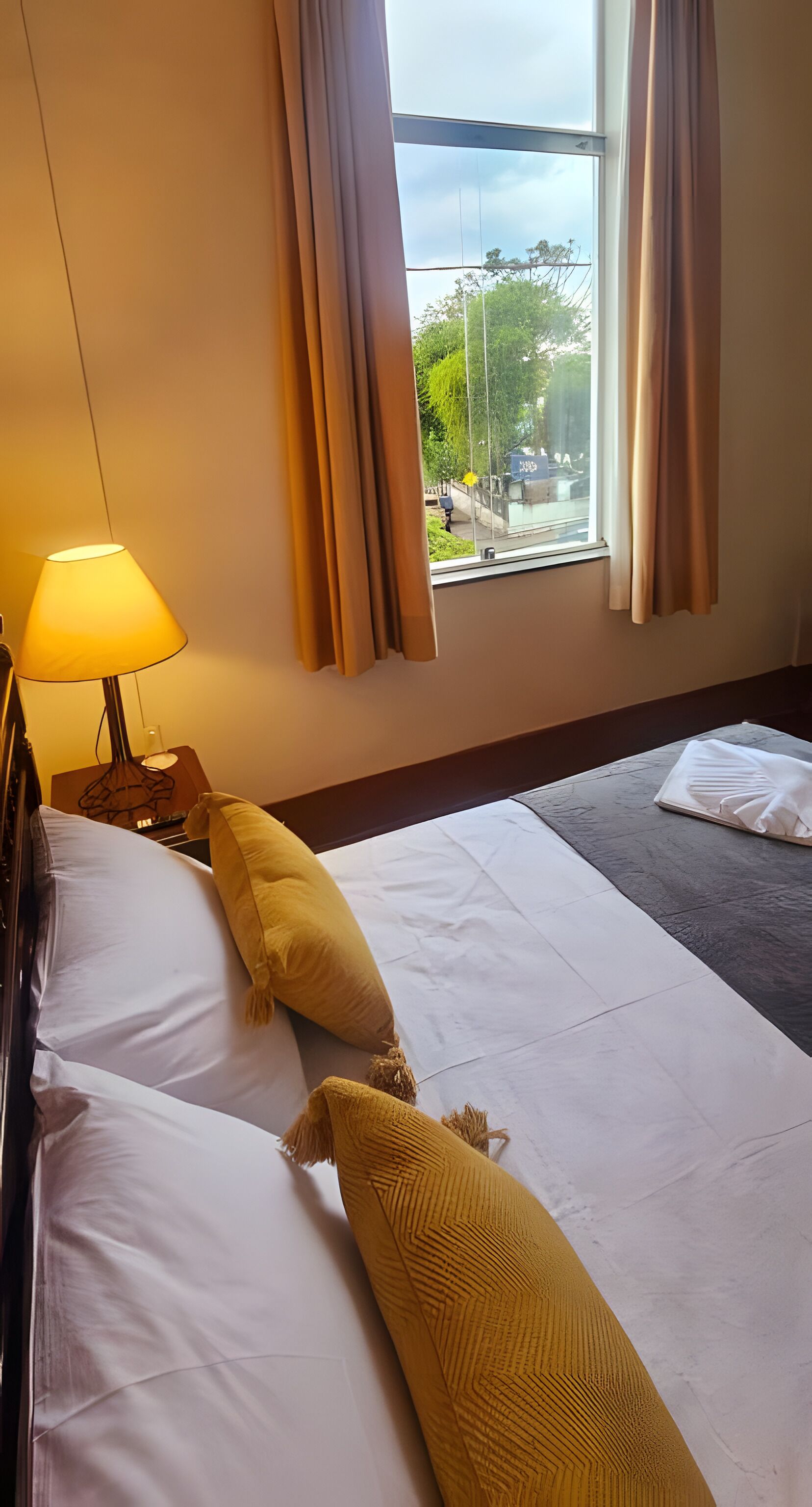 A hotel room with a bed, a lamp, and a view of the outdoors.