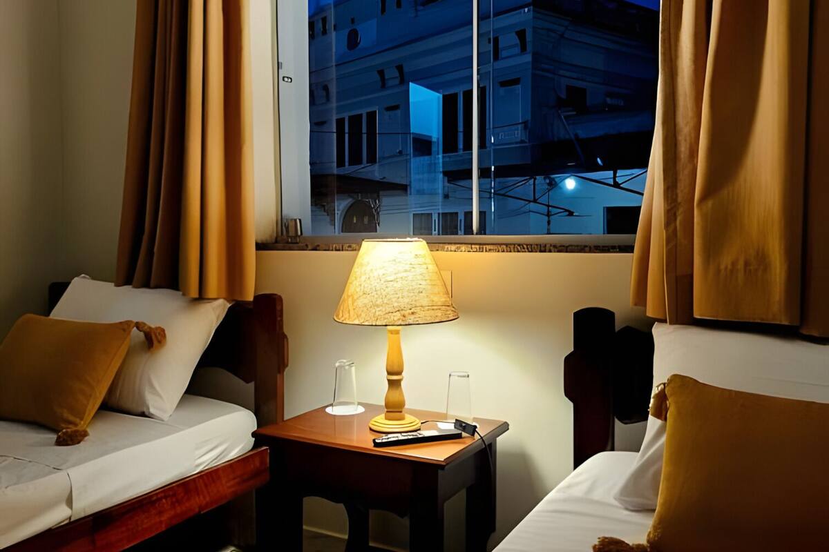 A hotel room with two beds, a nightstand with a lamp, a window with curtains, and a view of a building outside.