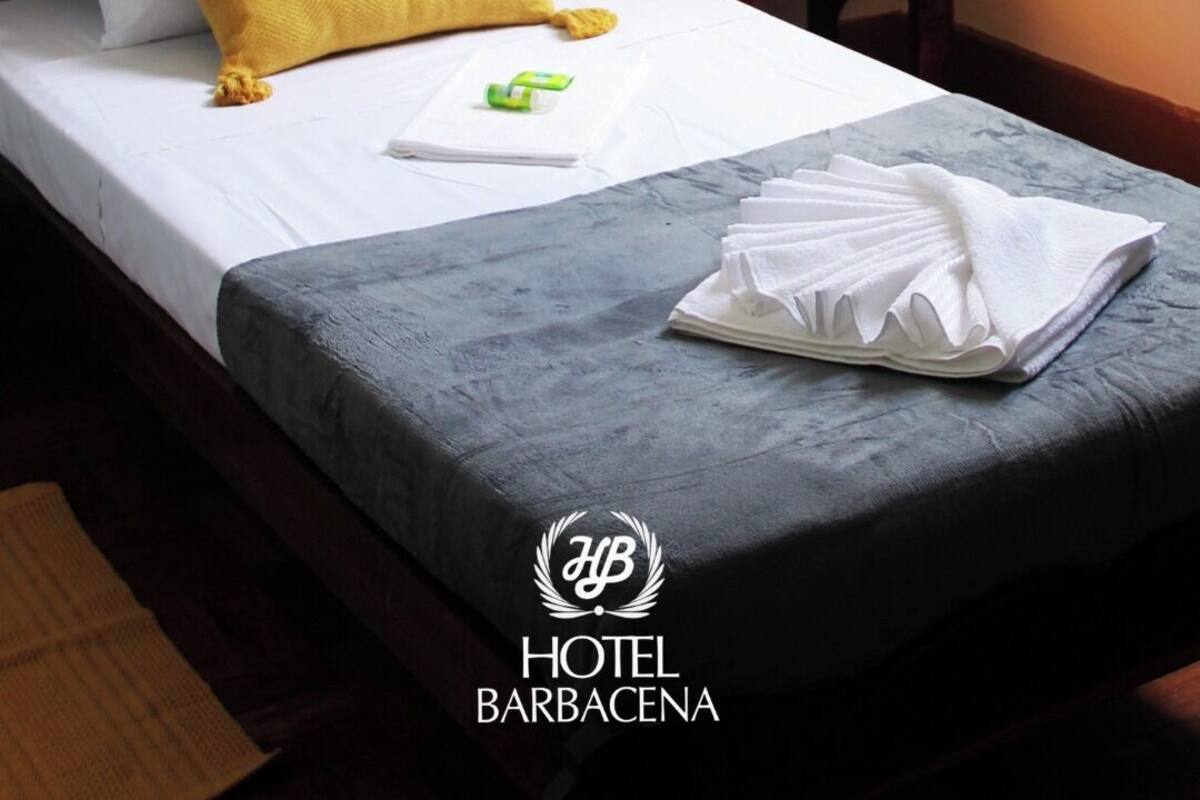 A neatly made bed with a white and yellow pillow, a folded white towel, and a small green item on the bed. There is a wooden nightstand with a lamp and a remote control beside the bed. The room has a simple, clean design with a visible logo of HOTEL BARBACENA.