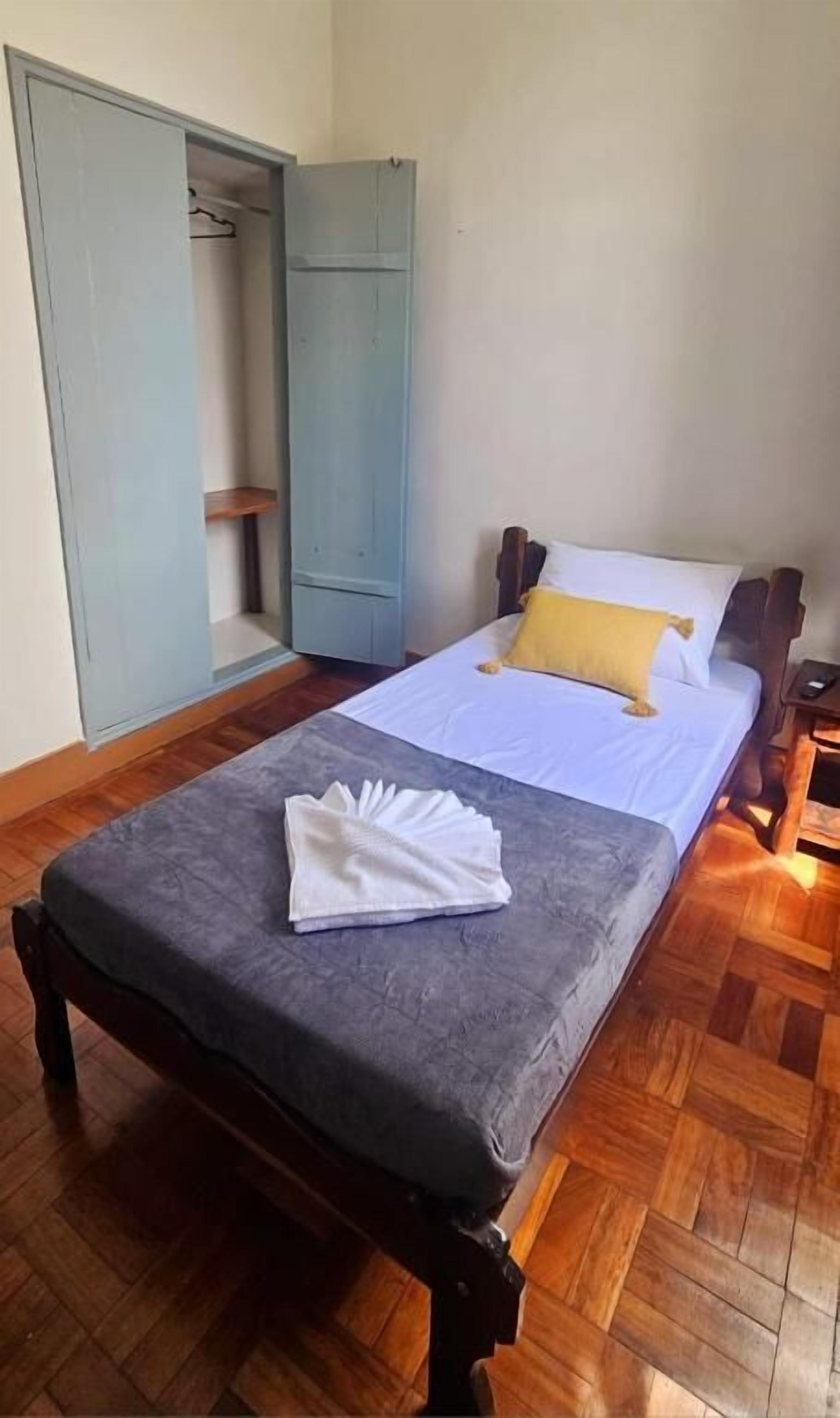 Uma cama de solteiro com um edredom cinzento, um travesseiro amarelo e uma toalha branca em assoalho de madeira.