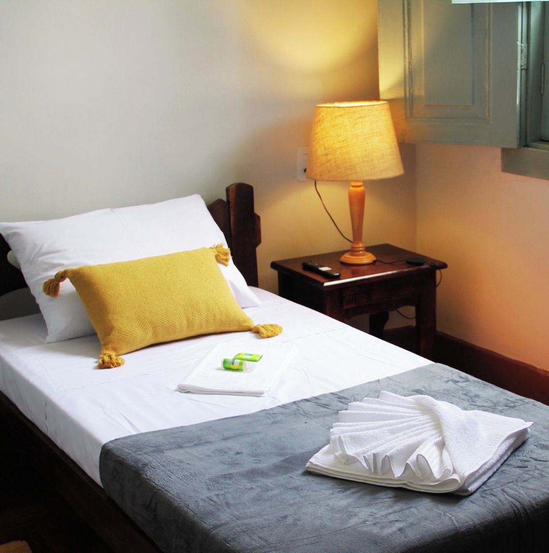 A neatly made bed with a white and yellow pillow, a folded white towel, and a small green item on the bed. There is a wooden nightstand with a lamp and a remote control beside the bed. The room has a simple, clean design with a visible logo of HOTEL BARBACENA.
