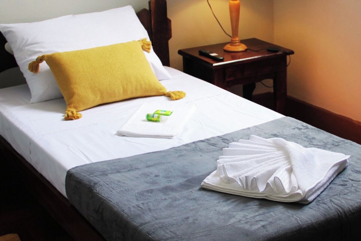 A neatly made bed with a white and yellow pillow, a folded white towel, and a small green item on the bed. There is a wooden nightstand with a lamp and a remote control beside the bed. The room has a simple, clean design with a visible logo of HOTEL BARBACENA.