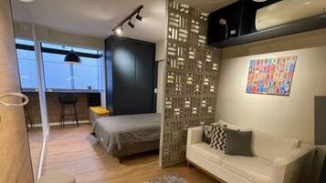 Quarto moderno com cama, sofá, escrivaninha e cadeira. Janela com vista para a cidade.