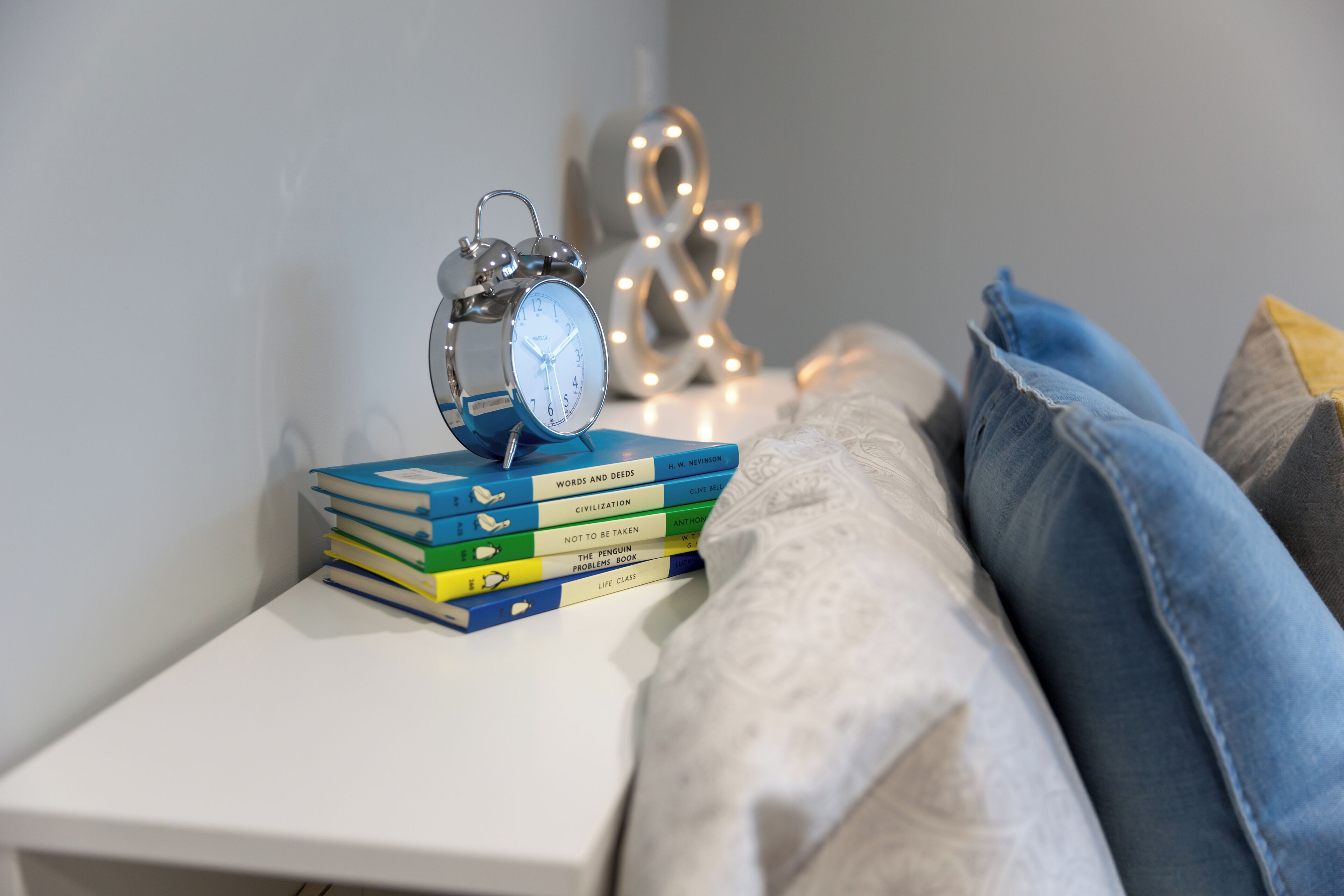 A bedside table with a stack of books, a silver alarm clock, and decorative lights.