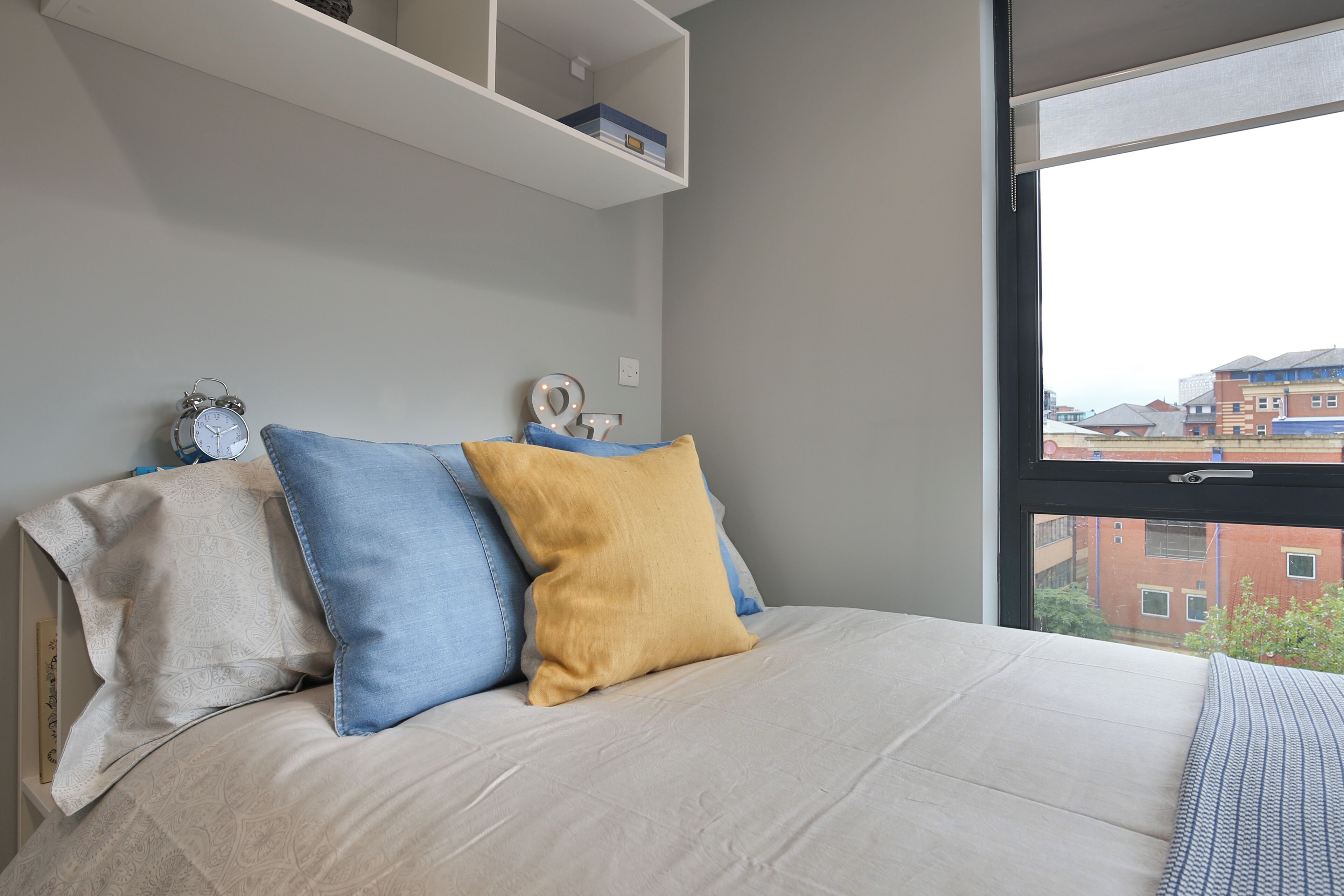 Une chambre à coucher avec un lit, des oreillers, une étagère et une fenêtre donnant sur des immeubles.