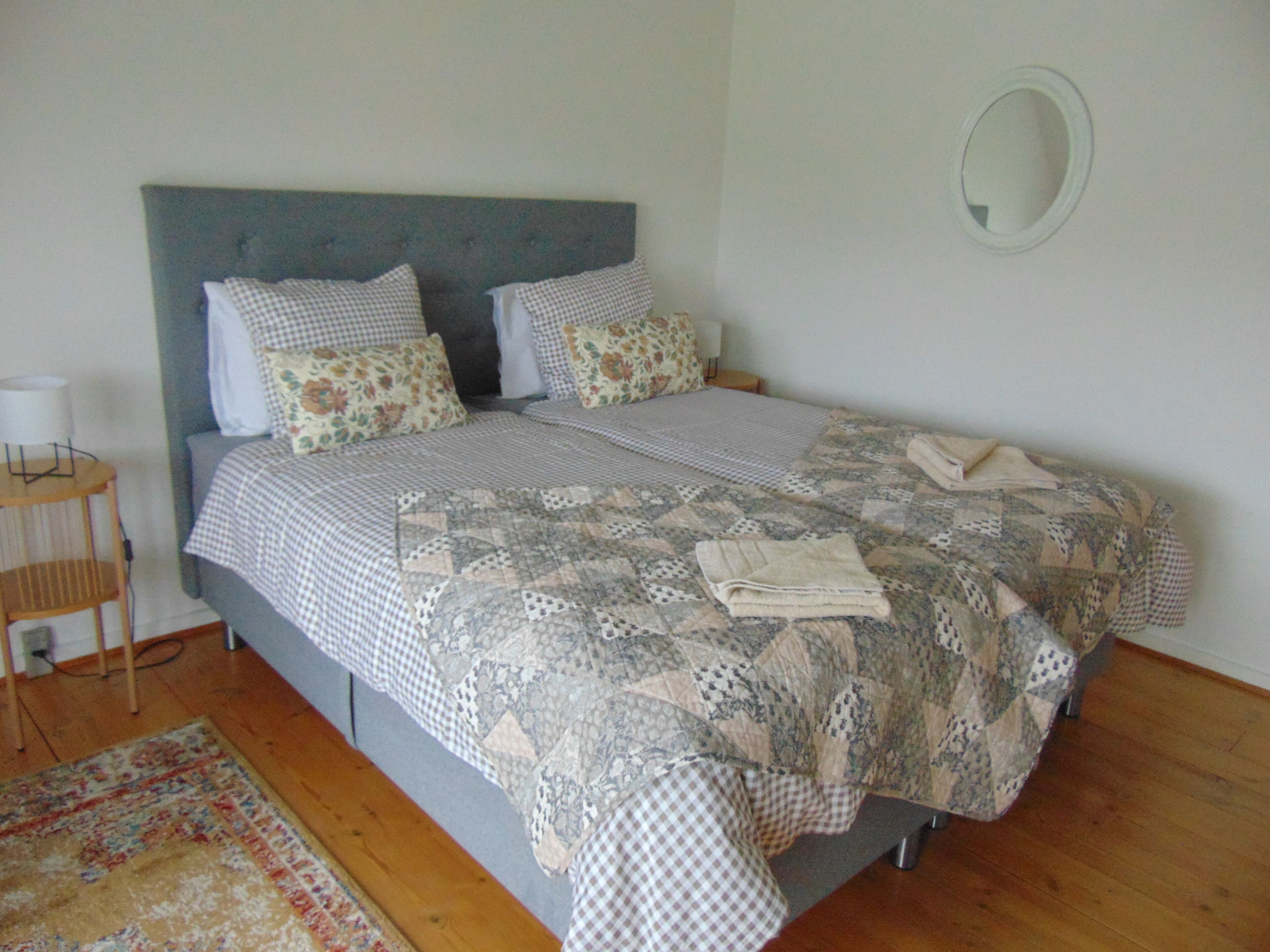 A neatly made bed with a checkered bedspread and floral pillows, a bedside table with a lamp, and a round mirror on the wall.