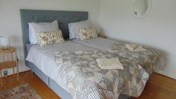 Una cama bien hecha con colcha a cuadros y almohadas florales, una mesita de noche con lámpara y un espejo redondo en la pared.