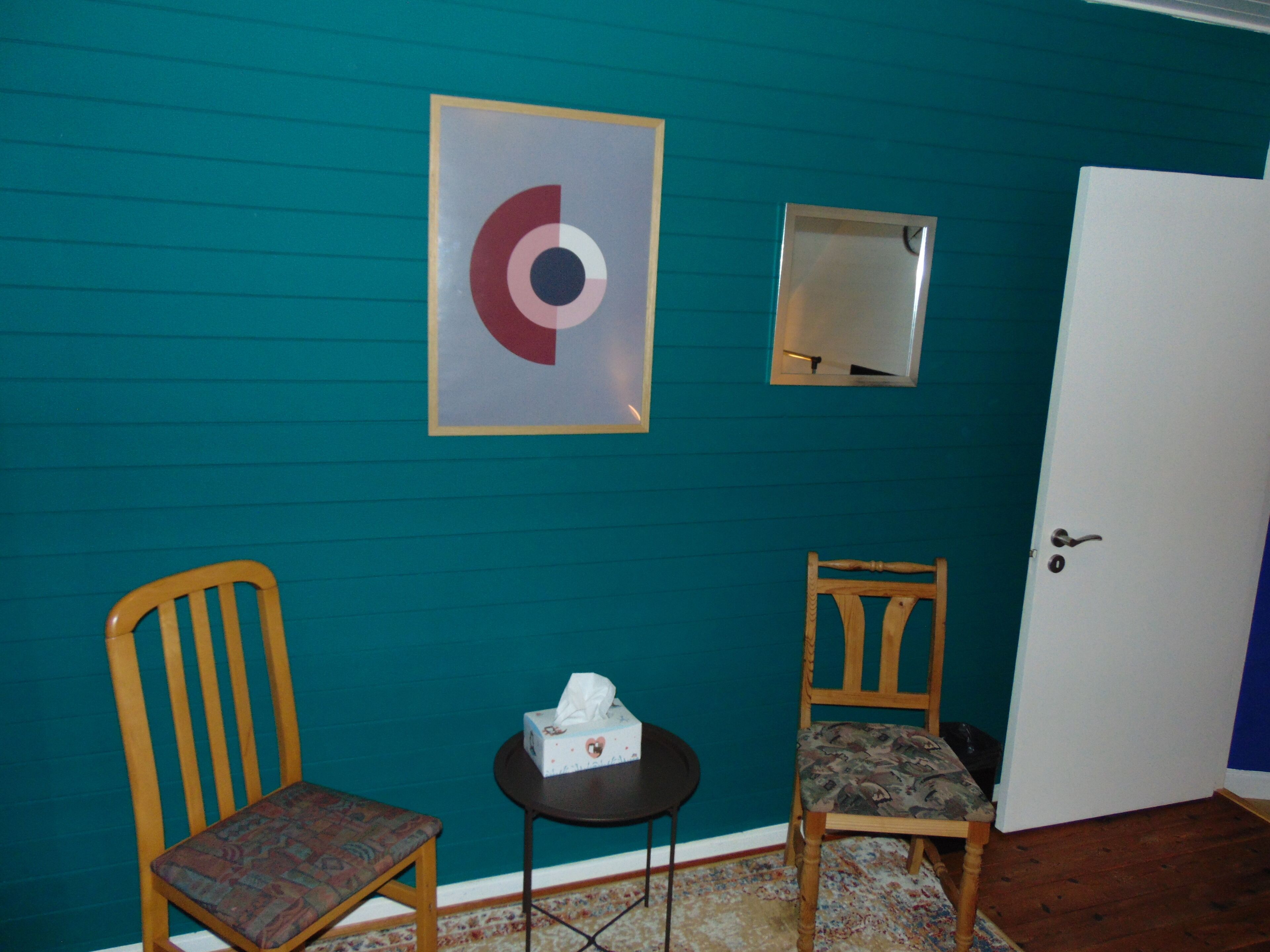 A teal-walled room with two wooden chairs, a small round table with a tissue box, and a framed abstract artwork.