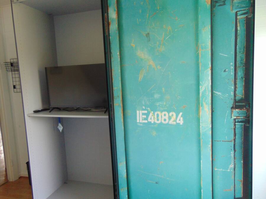 A room with a teal door marked with 'IE40824', a television on a shelf, and a small trash bin.