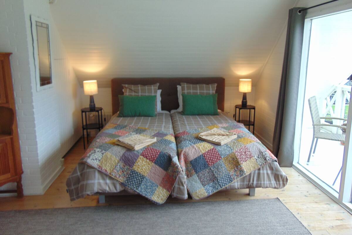 A bedroom with two beds, each with a quilt and pillows, a nightstand with a lamp, and a view of the outdoors through a large window.