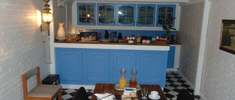Een eetgedeelte met een geruit vloertje, een houten tafel gedekt voor twee personen, en een keuken met blauwe kastjes en een wit aanrecht.