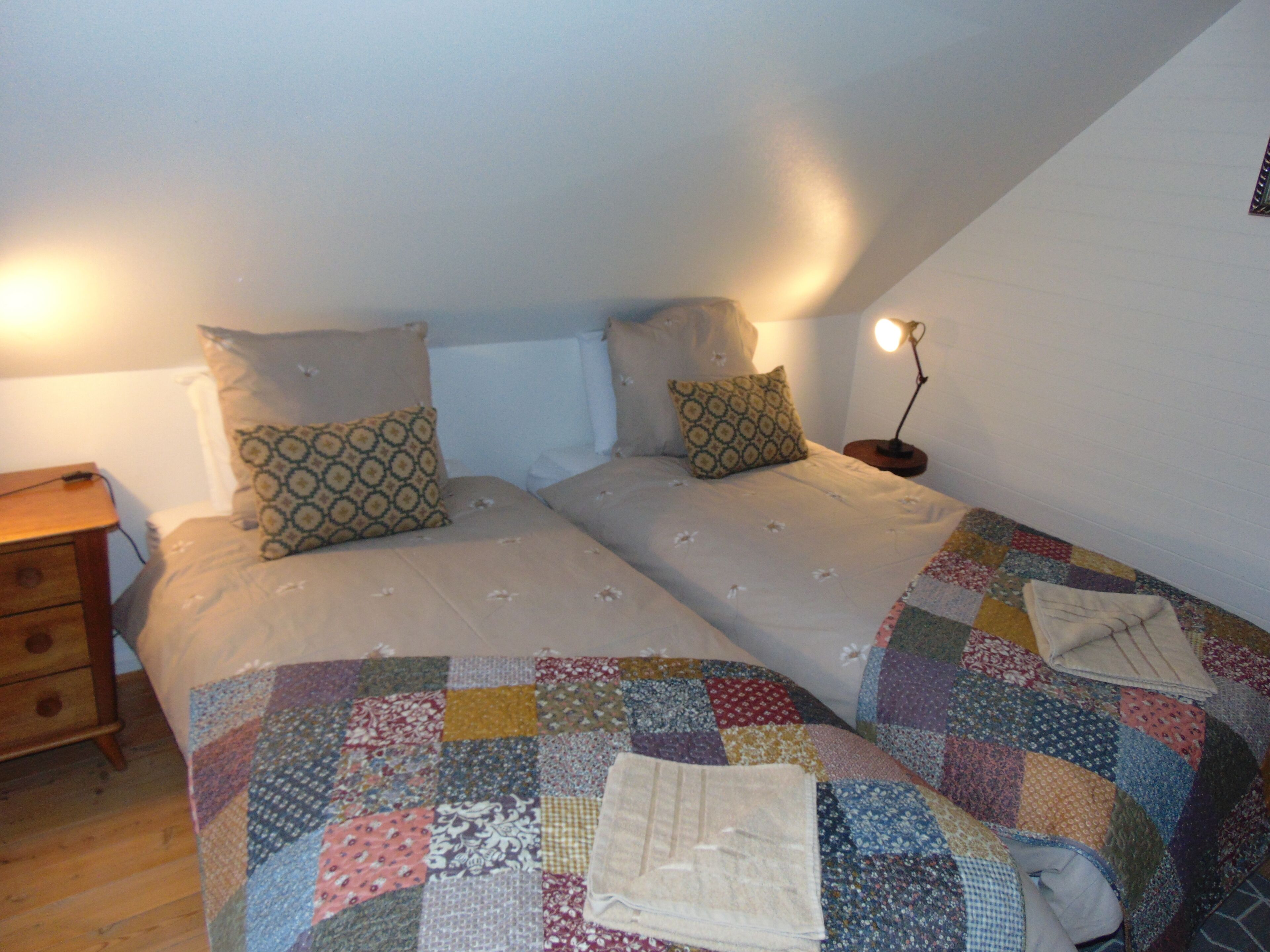 Un dormitorio con dos camas, una mesita de noche de madera, una lámpara y un colcha en el suelo.