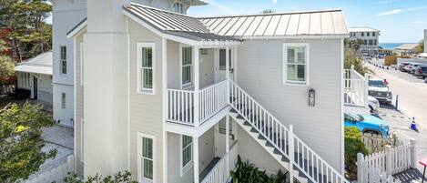 A multi-story white building with a central staircase, a covered porch, and a view of the beach.