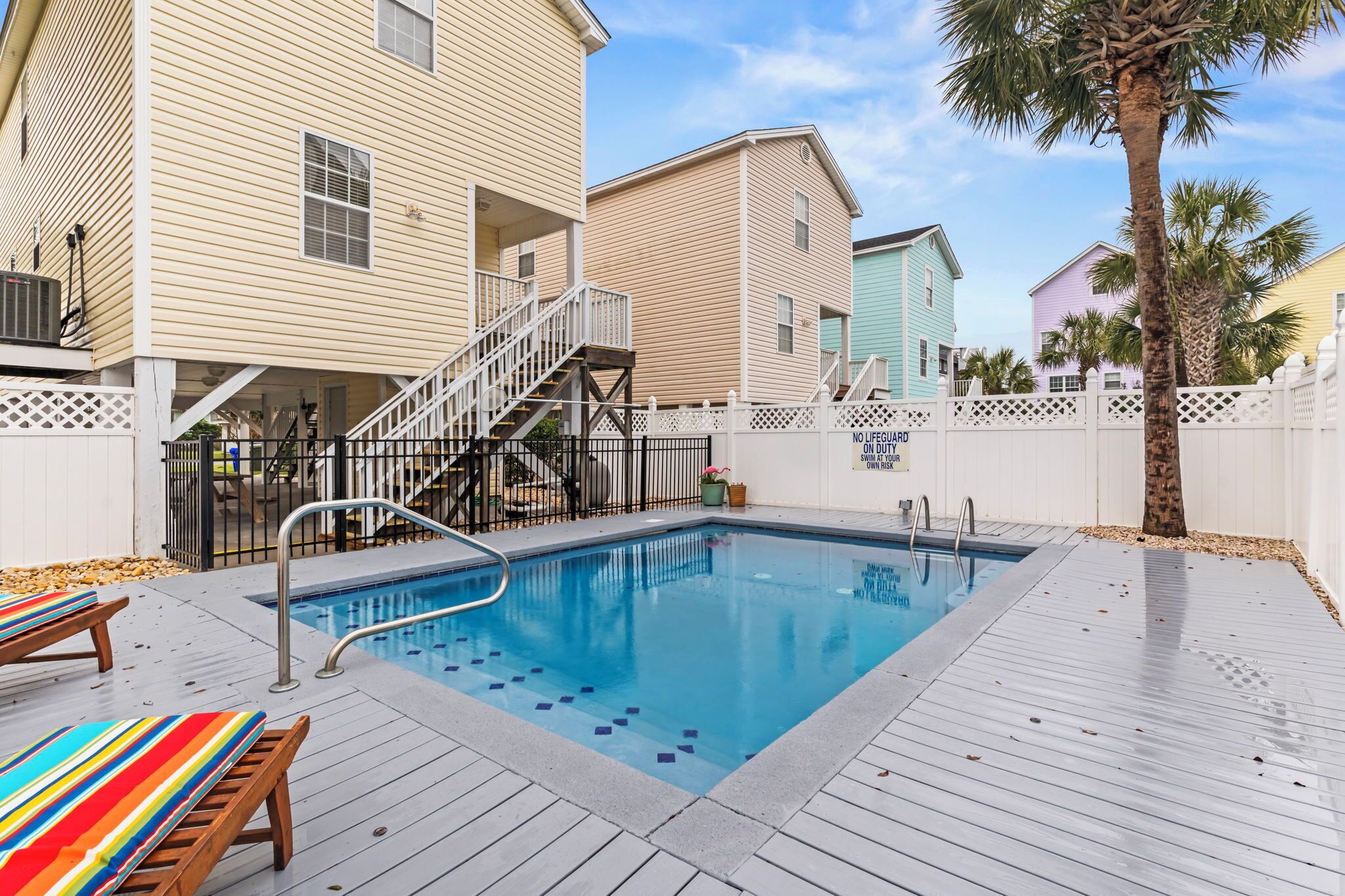 A swimming pool with a wooden deck, a white fence, and a staircase leading to a building.
