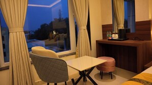 Ein Hotelzimmer mit großem Fenster, einer Couch, einem kleinen Tisch, einem Sessel, einem Schreibtisch und einem Spiegel.