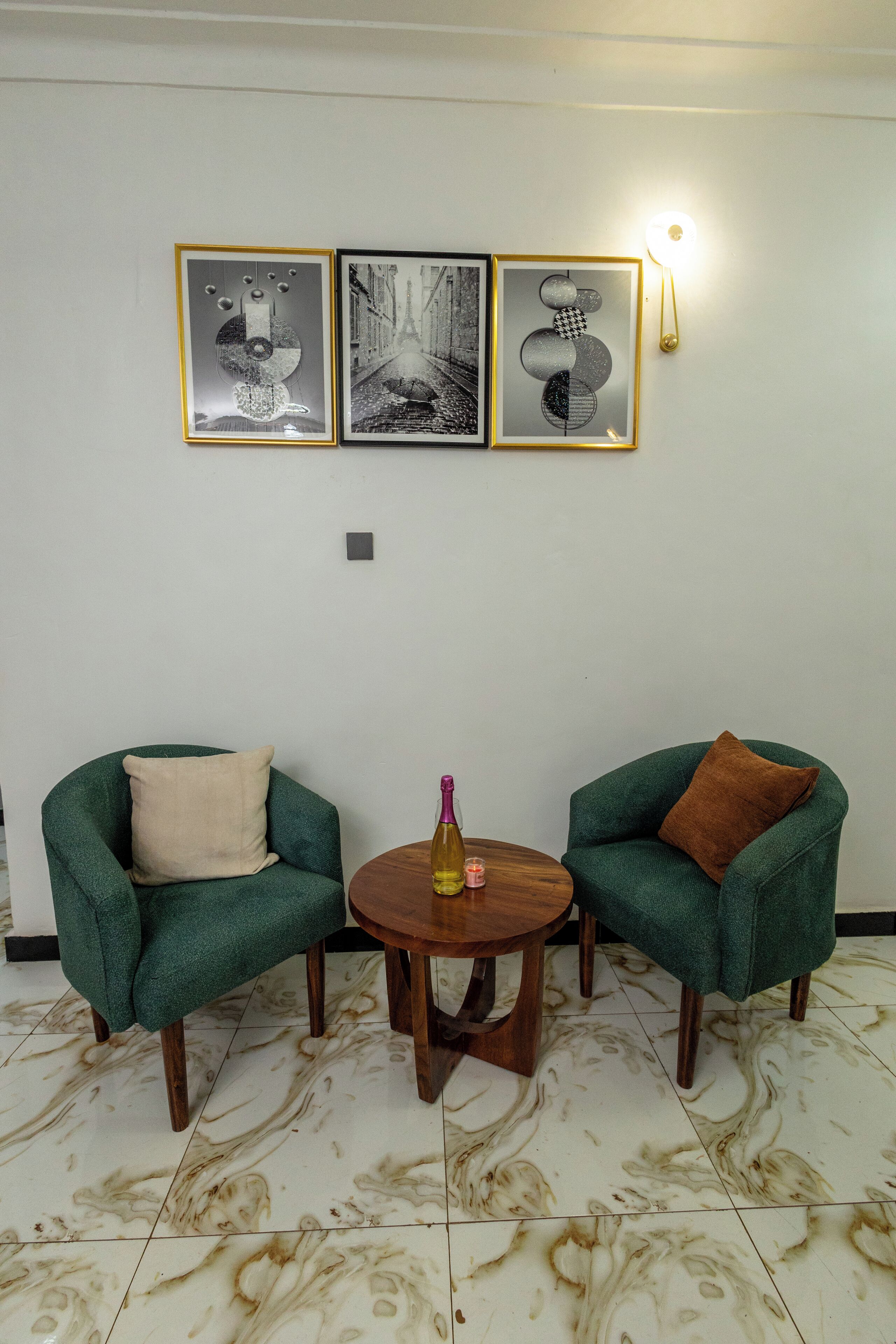 Una habitación con dos sillones verdes, una mesita de madera con una botella, y tres obras de arte enmarcadas en la pared.
